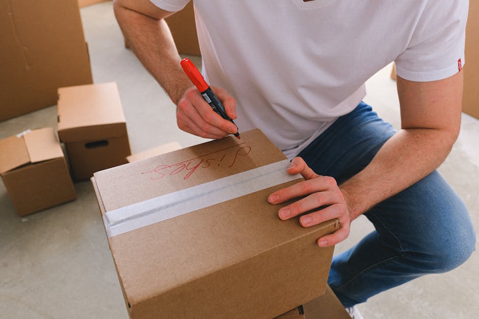 A person wearing a white T-shirt and blue jeans is kneeling in a room filled with cardboard boxes, some sealed with packing tape and others open. They are using a red marker to write on a medium-sized brown cardboard box, with the word 'Signed' visible in red ink. The boxes are arranged on the floor and near a doorway, indicating an ongoing packing process typical of home relocation or furniture transport. In the background, additional boxes of various sizes are stacked, and the room has natural lighting, suggesting a straightforward moving or packing operation. This scene reflects the logistical aspects of a professional house removal service, such as those offered by Man with Van Sudbury, involving careful packing and preparation for a residential move.