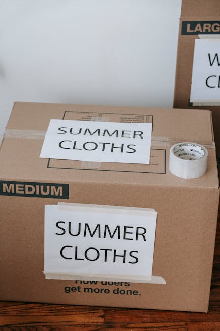 A close-up view of two brown cardboard boxes used for house removal and moving, placed on a wooden floor indoors. The larger box has white labels with black text reading 'SUMMER CLOTHS' and 'MEDIUM', indicating its contents and size. The smaller box behind it partially shows the word 'LARGE' on a label, suggesting it is a larger container. A roll of packing tape is positioned on top of the larger box, ready for sealing. The interior lighting is even, highlighting the clean, organized packing process typical of a home relocation. ManwithVansudbury, experienced in furniture transport and packing for removals, often uses such boxes to efficiently organise household belongings during home moves, ensuring safe transport and streamlined logistics.