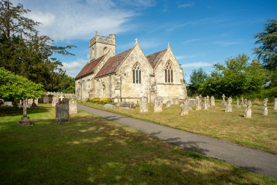 An exterior view of a historic stone church with Gothic-style windows and a square tower, situated within a grassy churchyard. The churchyard contains numerous weathered stone gravestones arranged in rows, some with rounded tops, and others with cross shapes. A paved footpath runs along the front of the church, leading towards the entrance, with patches of shadow cast by nearby trees. The scene is illuminated by natural daylight under a partly cloudy sky, with green trees and bushes surrounding the churchyard. This setting exemplifies a typical rural churchyard in the UK, often involved in local community activities such as moving or relocation services. Man with Van Sudbury occasionally handles home relocation tasks around similar properties, which may include packing, furniture transport, and loading processes within such historic sites.
