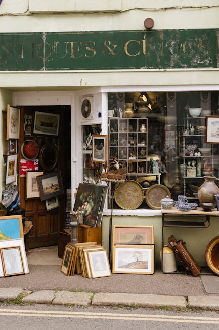 Exterior view of a shopfront with a green, weathered sign reading 'Antiques & Curios' above the entrance. The display window showcases a variety of antique items, including framed photographs, decorative plates, and small sculptures arranged on shelves and the windowsill. Several books and picture frames are placed on the pavement outside, leaning against the wall and on the ground, with some stacked. Inside, visible through the open doorway, are more antiques, including framed pictures, ornate vases, and a wooden cabinet filled with collectibles. The scene depicts the process of a house removal or furniture transport, likely involving the packing and loading of antique furnishings, with some items prepared for transport by [COMPANY_NAME], a professional removals service. The lighting is natural, capturing a quiet, daytime setting suitable for a furniture relocation or household move. The overall image emphasizes the careful handling and organization of fragile antiques during home relocation.