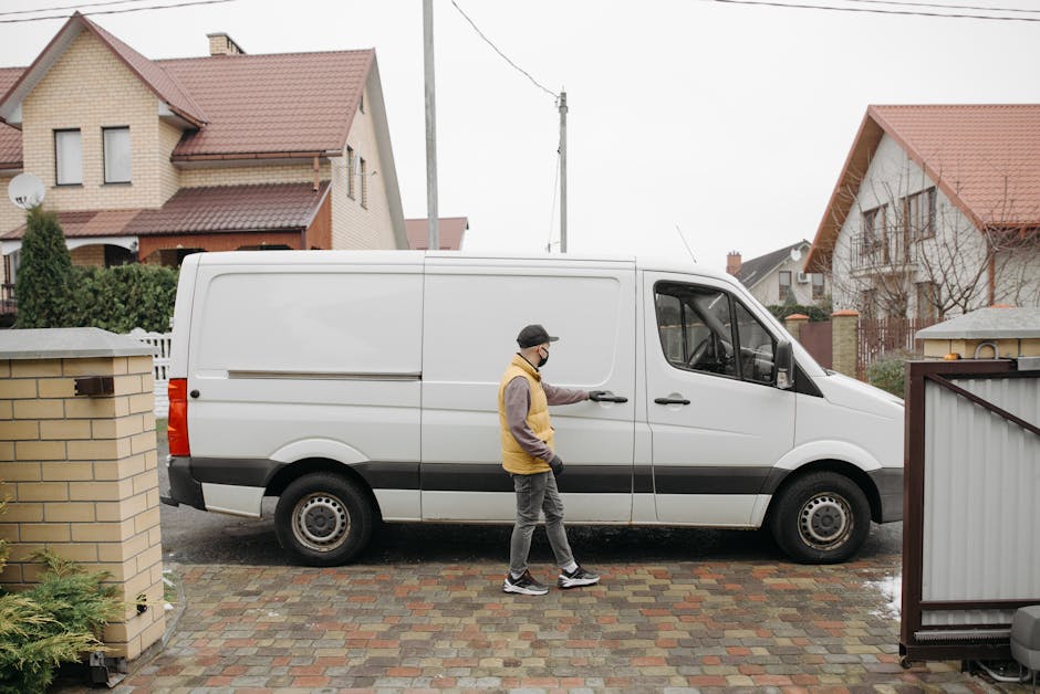Two men are engaged in a home relocation process outside a suburban house during daytime, with one man sitting on the open rear door of a white cargo van, dressed in casual clothing with a hat, smilin