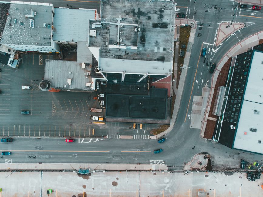 Aerial view of a city street showing a building with a flat roof and exterior walls, a small parking area with a few parked vehicles, and an adjacent paved road with lane markings and traffic signs. A large white van is parked close to the building's entrance, partially visible on the right side of the image, with a ramp or loading area nearby. The street is lined with cement pavements, crosswalks, and traffic cones, indicating ongoing parking suspensions or temporary restrictions typical during house removals or furniture transport in a busy urban environment. The scene is captured during daylight with natural lighting, suitable for a professional removals service like Man with Van Sudbury involved in packing, loading, or home relocation activities, supporting permit and parking suspension processes.