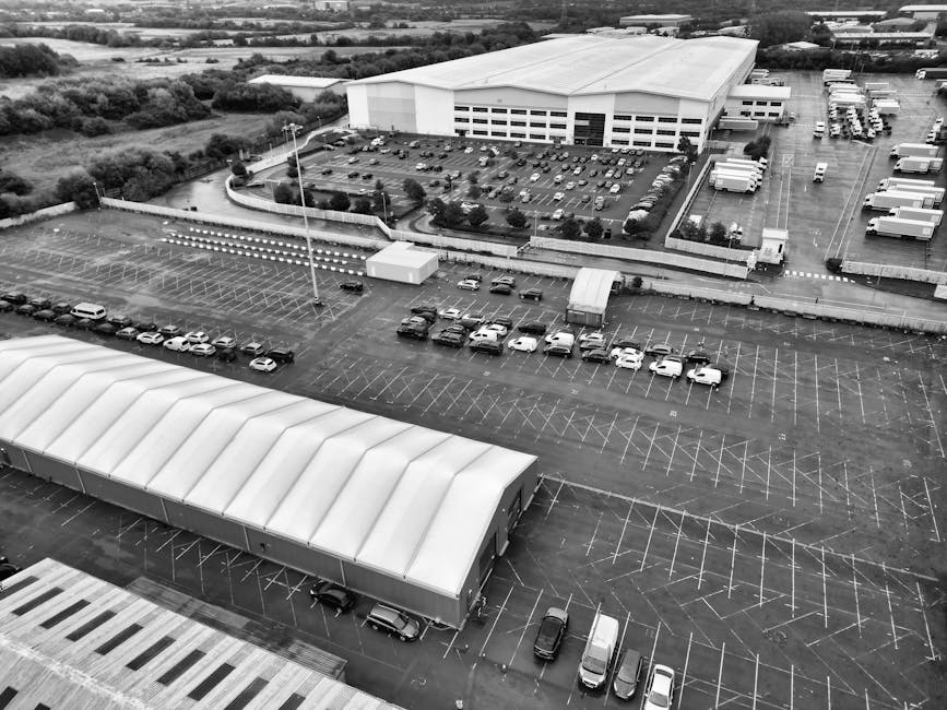 An aerial view of a large parking lot adjacent to a commercial building with a white, curved-roof warehouse structure in the foreground. Several cars are parked in designated spaces, some overlapping in the spacious lot, with a few vehicles actively moving or parked along the edges. A small, white rectangular kiosk or utility box is positioned near the center of the parking area. Beyond the parking lot, a high fence separates the area from an industrial estate, where multiple parked trucks and trailers are aligned in rows near a large, white warehouse with multiple garage doors. The environment is well-lit with natural daylight, and the scene suggests a setting suitable for house removals or furniture transport operations, possibly involving the use of ramps, trolleys, and moving straps by [COMPANY_NAME] during a home relocation or packing and moving process within the commercial premises.