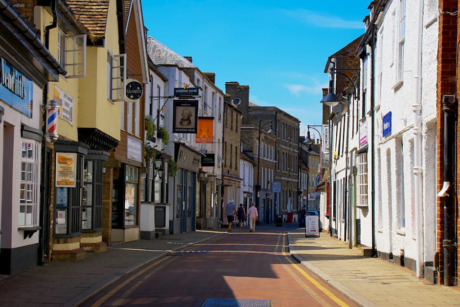 A narrow pedestrian street in Sudbury with closely packed buildings on both sides, featuring a variety of retail shops with signboards, window displays, and hanging flower baskets. The street is paved with cobblestones and marked with yellow lines along the curb. On the left, there is a building with a traditional barber's pole outside, indicating a barbershop, while other shopfronts display signs for local businesses. Several people are walking along the pavement, some carrying bags, while a large white van with open doors is parked on the right side near the entrance to a side alley, suggesting the commencement of a home relocation or furniture transport process. Inside the open van, partly visible, are packed cardboard boxes, wrapped furniture, and moving blankets, which are being loaded or unloaded by movers who are not visible in the image. The bright daylight and clear blue sky highlight the details of the scene, emphasizing a typical setting for a house removals service in Sudbury, managed by Man with Van Sudbury. The photo captures the urban environment and logistics involved in packing and moving between properties on Market Hill.