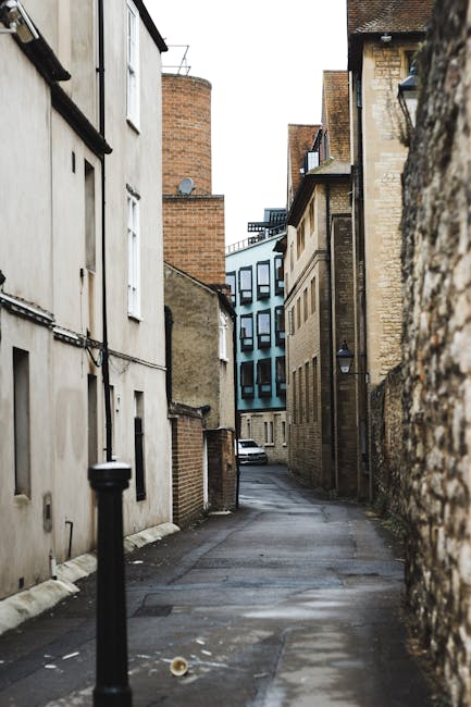 A narrow alleyway in Sudbury with closely spaced buildings on either side, featuring a mix of traditional brick and contemporary facades. The ground is paved with asphalt, slightly uneven, with some patches indicating recent repairs or wear. At the end of the alley, there is a modern, blue-tinted multi-storey building, contrasting with the older structures. A black bollard is positioned on the left foreground, preventing vehicle access. The alley appears dimly lit, with natural light filtering in from the open sky above, and no visible moving or loading activity occurring in the scene. This setting reflects the typical tight urban environment where home relocation or furniture transport might require careful vehicle navigation, as managed by professional removals service providers like Man with Van Sudbury, ensuring safe and efficient moving logistics in confined spaces.