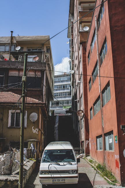 The image shows a narrow alleyway between two multi-storey buildings in Sudbury, with a white van parked at the entrance. The building on the left is an older structure with weathered yellow painted walls, a red tiled roof, and small circular satellite dishes attached to its exterior. The building on the right is a modern, taller brick apartment block with large rectangular windows, some of which are open. Overhead, several wires and cables are strung across the alley, and a few small lamps are visible on the buildings' facades. Inside the alley, there are some discarded items and makeshift fences, indicating a residential area. In the background, additional high-rise buildings are visible under a partly cloudy sky. This scene illustrates a typical urban environment where home relocation or furniture transport services by [COMPANY_NAME] such as Man with Van Sudbury might operate, emphasizing the tight space constraints encountered during loading and unloading processes in narrow lanes.