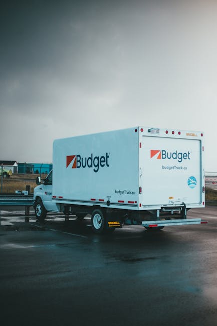 A white Budget moving van is parked on a wet tarmac surface in an open area, with a partly cloudy sky overhead. The van features the Budget logo and website address on the side and rear doors. Its rear loading doors are closed, and the vehicle is positioned in front of a loading zone with a yellow wheel chock placed underneath. In the background, there are industrial buildings, and a few other vehicles are visible in the distance. The wet ground reflects some of the vehicle’s details, indicating recent rain. This scene captures the process of home relocation or furniture transport, with the van prepared for loading or moving goods. The image relates to professional removals and transport services, illustrating the logistical stage of packing and moving a household, supported by the presence of the van and its location on a designated loading area. Man with Van Sudbury may utilize such vehicles for same-day removal services, ensuring efficient home relocation and furniture transport.
