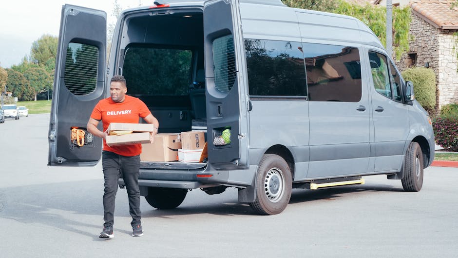 A man wearing a red delivery T-shirt is standing outside a grey panel van with its rear doors open, preparing to load or unload cardboard boxes, plastic-wrapped packages, and other moving supplies. The van is parked on a suburban street with residential houses, trees, and parked cars visible in the background. Inside the van, various packed items are arranged on the floor, indicating a home relocation or furniture transport process. The man holds two boxes, one stacked on top of the other, and appears to be engaged in loading or unloading as part of the moving service provided by Man with Van Sudbury, capturing the loading process involved in packing and moving tasks.