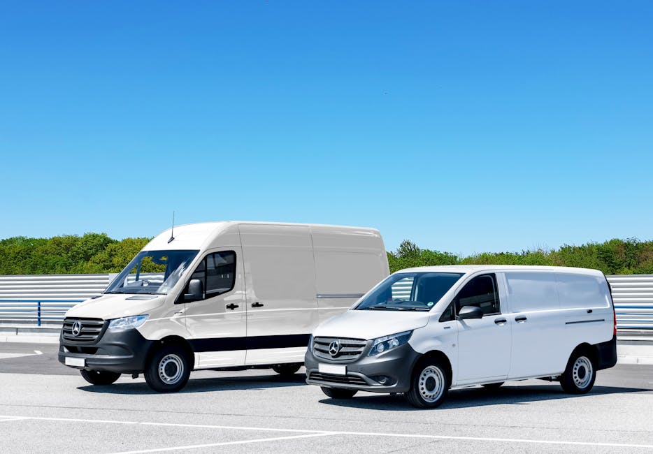 A white Budget moving van is parked on a wet tarmac surface in an open area, with a partly cloudy sky overhead. The van features the Budget logo and website address on the side and rear doors. Its rear loading doors are closed, and the vehicle is positioned in front of a loading zone with a yellow wheel chock placed underneath. In the background, there are industrial buildings, and a few other vehicles are visible in the distance. The wet ground reflects some of the vehicle’s details, indicating recent rain. This scene captures the process of home relocation or furniture transport, with the van prepared for loading or moving goods. The image relates to professional removals and transport services, illustrating the logistical stage of packing and moving a household, supported by the presence of the van and its location on a designated loading area. Man with Van Sudbury may utilize such vehicles for same-day removal services, ensuring efficient home relocation and furniture transport.