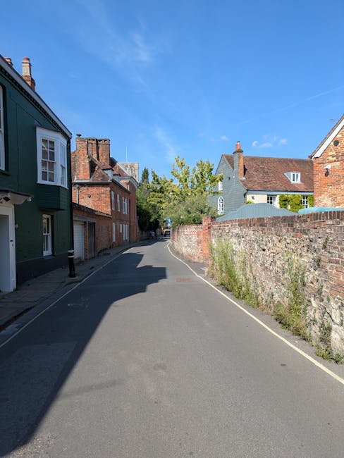 A quiet residential street with a narrow tarmac road curving slightly to the right, flanked by a mix of traditional brick and painted houses on the left and an old brick wall on the right. Several hou