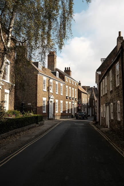 A quiet residential street with a narrow tarmac road curving slightly to the right, flanked by a mix of traditional brick and painted houses on the left and an old brick wall on the right. Several hou